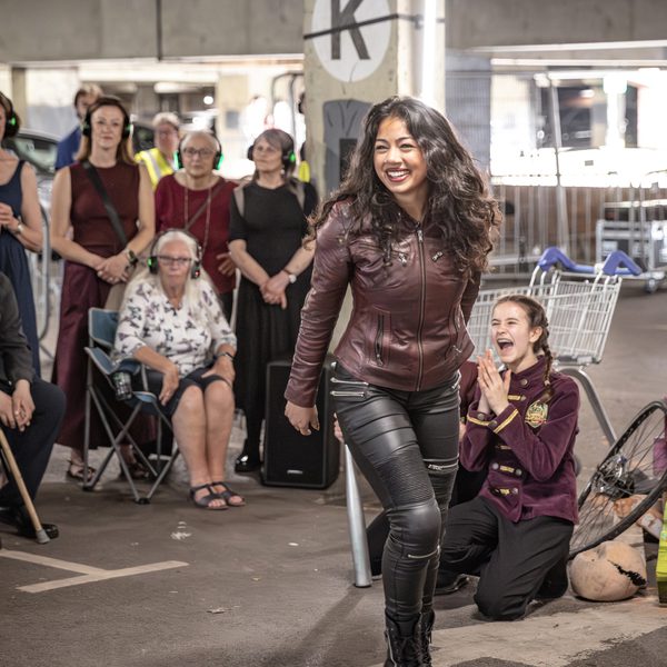 Actor Shaniya Hira performing as Tybalt in Wiltshire Creative's production of Romeo and Juliet. She's standing in a set built around a multi-storey car park, holding a baseball bat and grinning. Audience and seated and standing behind her wearing headphones.