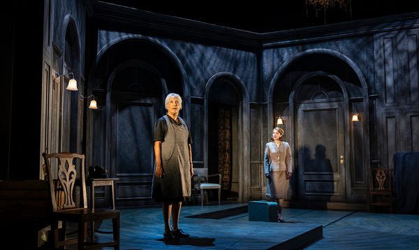 Two woman stand within a set built to look like the interior of an old house. The lighting is dark and atmospheric. One woman wears an apron, while another wears a suit jacket, with a bag at her feet, as if she's just arrived.