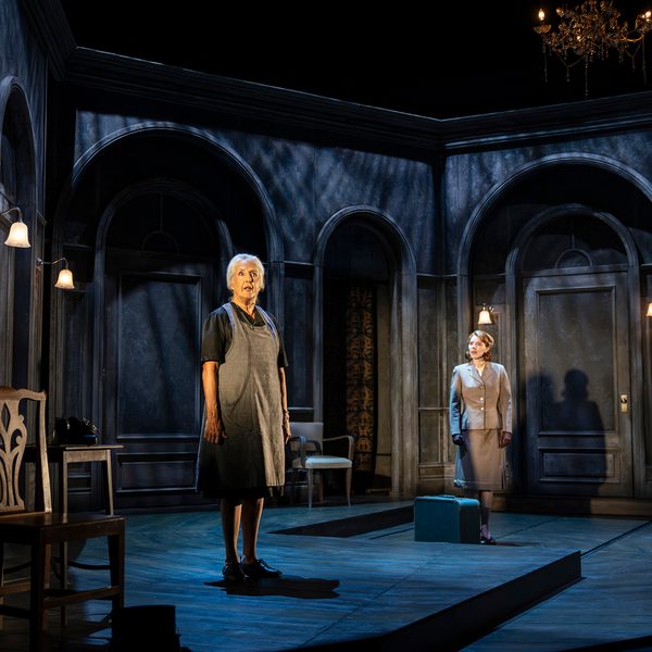 Two woman stand within a set built to look like the interior of an old house. The lighting is dark and atmospheric. One woman wears an apron, while another wears a suit jacket, with a bag at her feet, as if she's just arrived.
