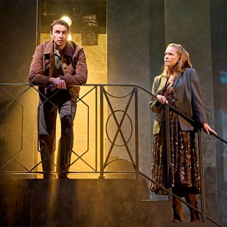 A production image from ‘Don’t Look Now.’ Laura stands half-way down some stairs, holding onto the railings, whilst John is at the top of them. Laura looks at John, worried. A glowing yellow light illuminates John from behind, from a doorway he is standing in front of.