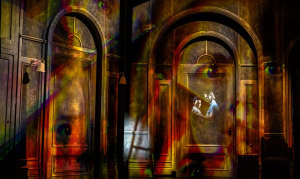 A man and a woman stand together. The man has a hand placed on the woman's upper arm. They are seen hazily through a window frame within the walls of a house. A large, fractured image of another woman's face is distorted across the walls.