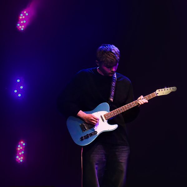 A young person playing a guitar on stage, with purple lights behind them.