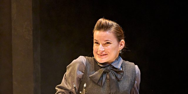 A production image from ‘Don’t Look Now.’ The hotel Clerk, wearing a smart black uniform, smiles as she stands behind a desk.