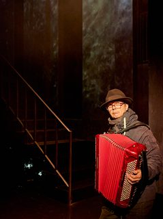 A production image from ‘Don’t Look Now.’ A man stands a few feet away from the bottom of a staircase and is playing a red accordion.