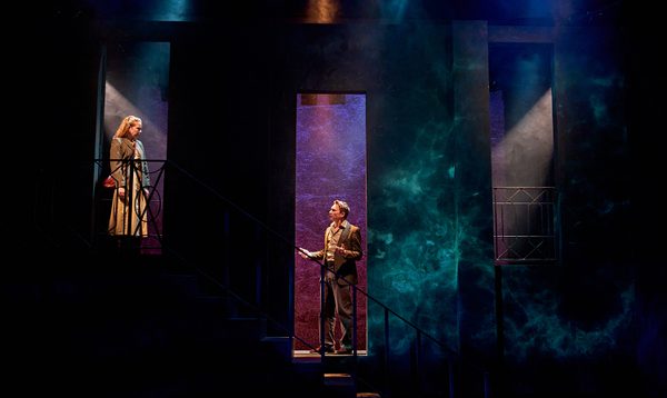 A production image from ‘Don’t Look Now.’ Laura stands at the top of some stairs, looking down at John who is stood half-way up the stairs. John is speaking, and is gesturing with both arms out either side of his body.