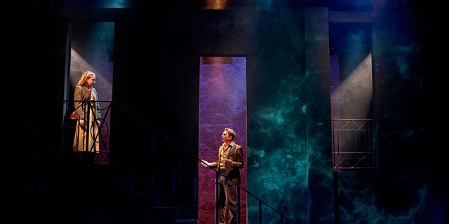 A production image from ‘Don’t Look Now.’ Laura stands at the top of some stairs, looking down at John who is stood half-way up the stairs. John is speaking, and is gesturing with both arms out either side of his body.