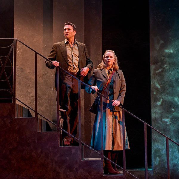 A production image from ‘Don’t Look Now.’ John and Laura stand half-way down some stairs, holding onto the railings. They both have slightly stunned looks on their faces, as if they have been spooked by a sight or a sound.