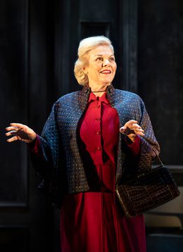A woman wearing a jacket over a dress and holding a handbag on her forearm. She is gesturing out with her hands and smiling.
