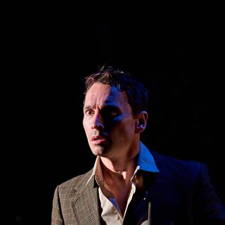 A production image from ‘Don’t Look Now.’ John, wearing a smart suit, looks at something off-camera, with a wide-eyed, shocked expression on his face, Only half of his face is lit by the stage lighting.