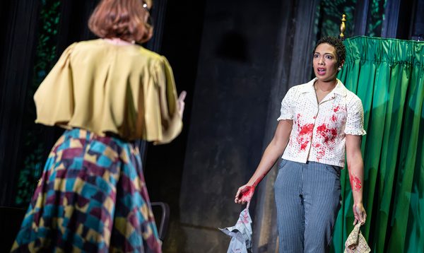 A woman faces the camera, dangling garments from her hand. Her shirt and forearms are marked with blood and she has a horrified expression on her face. Another person faces her, with their back to the camera.