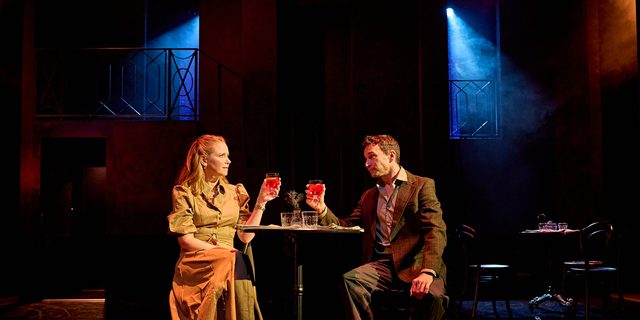 A production image from ‘Don’t Look Now.’ Laura and John are sat at a table in a restaurant. They are both holding up their glasses, about to clink them together.