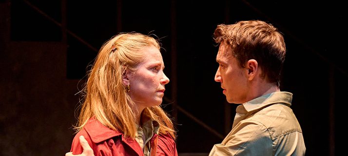 A production image from ‘Don’t Look Now.’ John and Laura stand together. Laura is holding a red, leather travel bag. John, standing in front of Laura, uses both hands to hold her shoulders, perhaps trying to stop her from leaving.