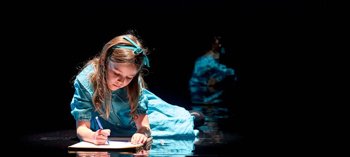 A production image from ‘Don’t Look Now.’ A young girl wearing a light blue dress is laying on the floor, drawing a picture on a piece of paper. He reflection can be seen on the shiny black floor.