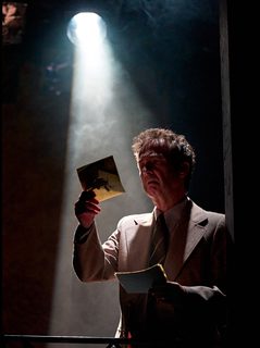 A production image from ‘Don’t Look Now.’ A man stands in a spotlight, looking concerned as he looks at a piece of paper which he holds in his right hand.