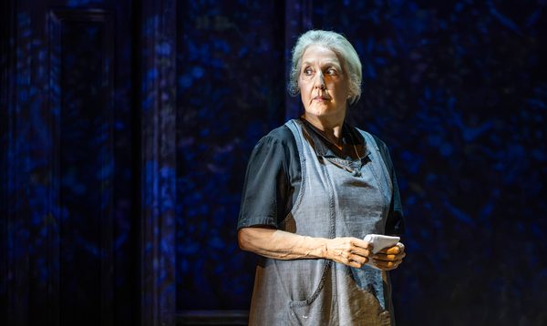 A woman wearing an apron, holding a letter, looking into the distance. She stands alone against a dark backdrop with partial, fractured lighting.