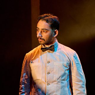 A production image from ‘Don’t Look Now.’ A restaurant waiter stands, wearing a smart white shirt and black bow tie. He is listening intently to something being said by another character.