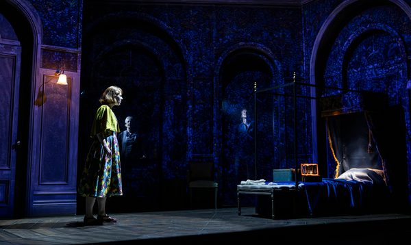 A woman stands staring at a four-poster bed. The scene is dark, lit with an eerie blue light, and there appears to be a shadow rising from the head of the bed.