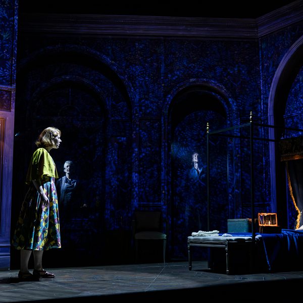 A woman stands staring at a four-poster bed. The scene is dark, lit with an eerie blue light, and there appears to be a shadow rising from the head of the bed.