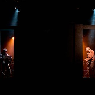 A production image from ‘Don’t Look Now.’ John, standing in a doorway on the right of the image, looks across to the other side of the stage, where another man is playing an accordion.