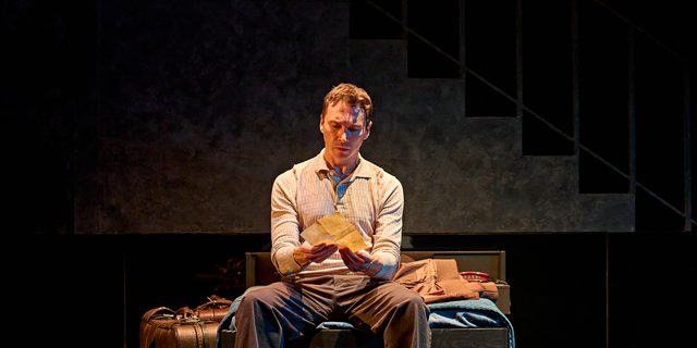 A production image from ‘Don’t Look Now.’ John is sat on the edge of a bed, reading a note which he holds in both hands.
