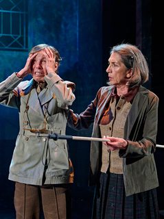 A production image from ‘Don’t Look Now.’ The two sisters stand together. Sister 1, on the right, places her hand on the shoulder of Sister 2, who is holding both hands to her head and has an extremely panicked expression on her face. The stage behind them is lit a deep blue colour.