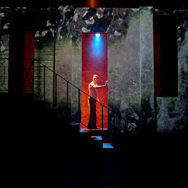 A production image from ‘Don’t Look Now.’ John, looking tired, stands in the centre of a doorway, resting his hand up against one side of it. The wall behind him is lit a deep red colour.