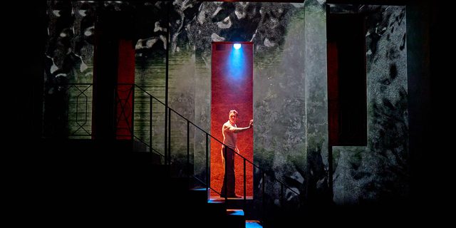 A production image from ‘Don’t Look Now.’ John, looking tired, stands in the centre of a doorway, resting his hand up against one side of it. The wall behind him is lit a deep red colour.