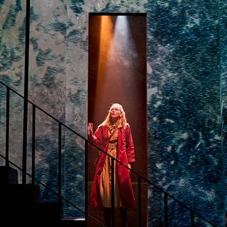 A production image from ‘Don’t Look Now.’ Laura stands in a doorway, with one hand resting on the door frame. She is looking up towards the sky, nervously. She is wearing a long, bright red coat.