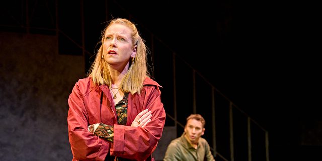 A production image from ‘Don’t Look Now.’ In the background, John is sat down on a bed, looking at Laura, in the foreground. Laura is standing, with her arms folded, and is looking at something off-camera with a concerned expression on her face.