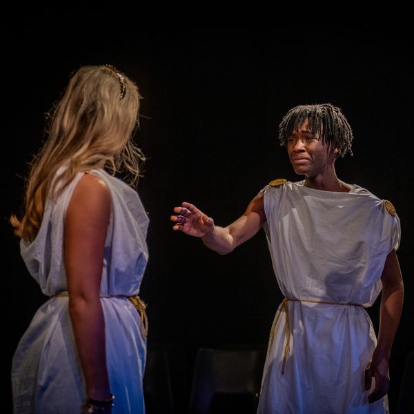 Two Wiltshire Creative Academy students performing in a play. They are both dressed in white robes. One of the young actors is reaching out to another who has their back to the camera.