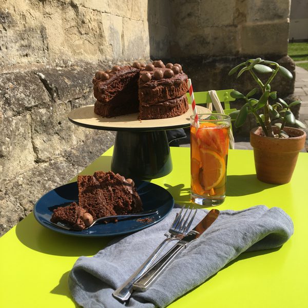 A photo of a chocolate cake, an orange drink, a plant pot, a plate with a cake slice and a knife and fork laid out on a green table in an outdoor space.