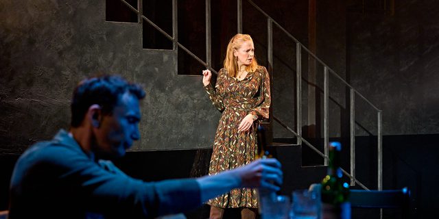 A production image from ‘Don’t Look Now.’ John, blurred in the foreground, is sat at a restaurant table, picking up a bottle. In the background, Laura stands next to some stairs. She has a confused look on her face, as if she has just been told something shocking.