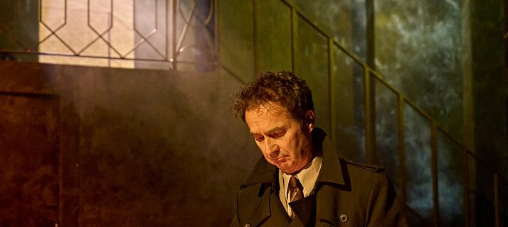 A production image from ‘Don’t Look Now.’ A man is standing behind a table with stacks of white candles on it. He has picked up one of them and is lighting it with a match.