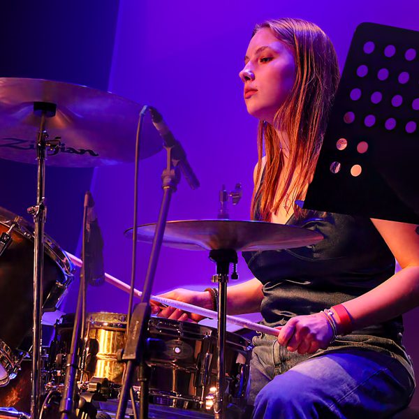 A young woman playing the drums, lit by purple light.