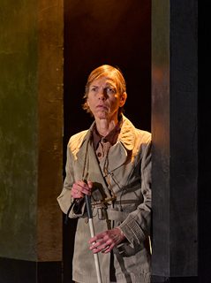 A production image from ‘Don’t Look Now.’ One of the sisters stands in a doorway, with a glowing yellow light shining from above her. She is holding a walking stick in her hands and is looking towards the centre of the stage, listening intently.