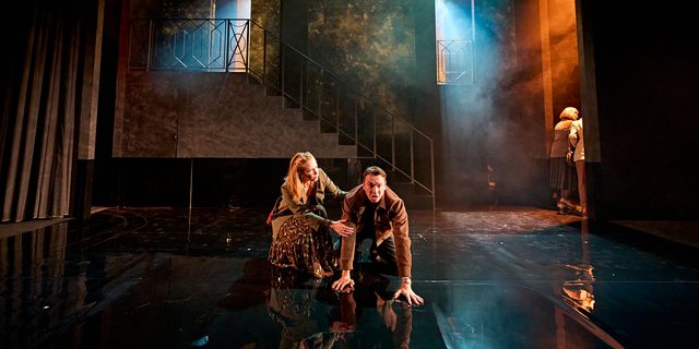 A production image from ‘Don’t Look Now.’ Laura kneels beside John, who is on his hands and knees in the centre of the stage. Laura looks to be comforting John, who has an angry/distraught expression on his face.