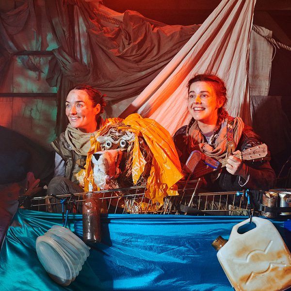 The two actresses from Stories in the Dust. They are sitting in a shopping trolley with a lion puppet in between them. They are both smiling off camera. One of them is playing a ukelele.