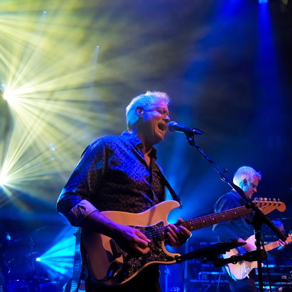 Two men playing guitar and singing on stage, surrounded by an impressive light show.
