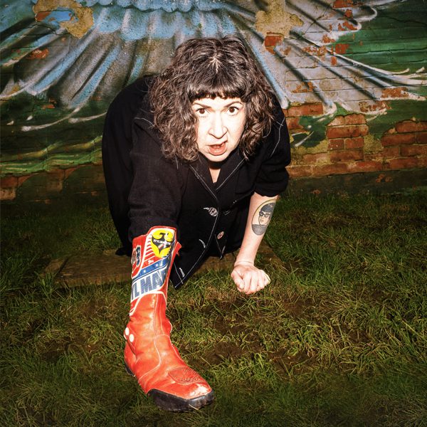 Comedian Amy Mason crouching towards the camera in a grassy field wearing orange boots.