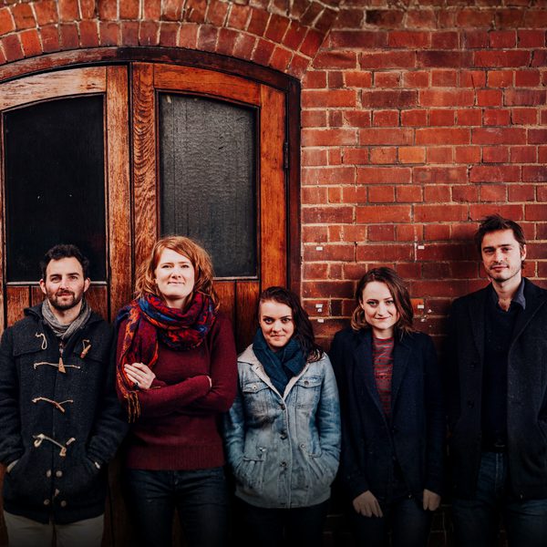 The three members of Lady Maisery, and folk duo Jimmy & Sid, all lined up in winter coats against a red-brick wall.