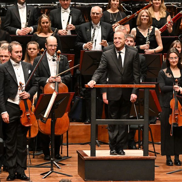 Mark Wigglesworth and the Bournemouth Symphony Orchestra dressed in formal attire, bowing after a concert in a large wooden hall.