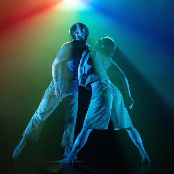 Two dancers (one male, one female) performing on stage together, washed by rainbow prism-like light.