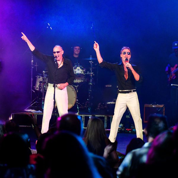 Two singers performing onstage, holding microphones with their right hands in the air.