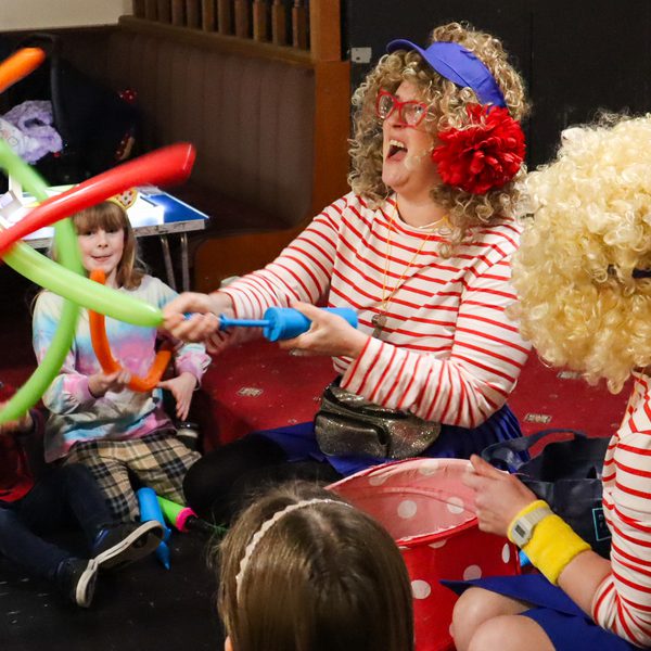 two performers wearing red and white stripes and curly wigs are playing with long thin balloons. Children are sat on the floor watching