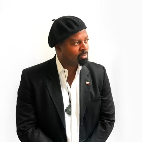 Poet Sir Ben Okri standing in a black beret and suit jacket against a white background.