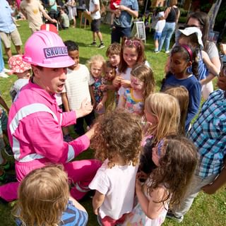 A performer dressed in pink hi-viz jacket and hard hat crouching down, surrounded by children