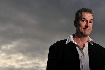 A photo of comedian Boothby Graffoe in front of a grey sky. He is smiling and wearing a shirt and suit jacket.