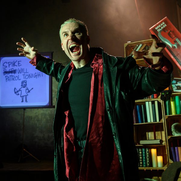 Comedian Brendan Murphy dressed as Spike from Buffy the Vampire Slayer performing on stage in front of a whiteboard and bookshelf. He is posing menacingly with his arms raised and holding an orange book.