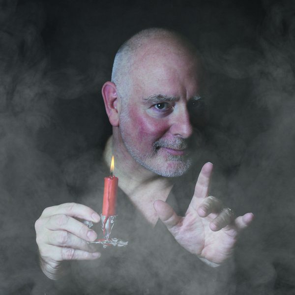 Actor Guy Masterson holding a red candle and posing spookily against a black smoky background.