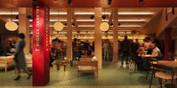 An artist's impression of the warmly lit interior of Salisbury Playhouse foyer, with wooden furniture, round pendant lights, and people seated at tables or walking through the space. A red column in the foreground displays white text reading “SALBERG STUDIO" and “MAIN THEATRE”, while the background shows a bar area and structural columns under a coffered ceiling.